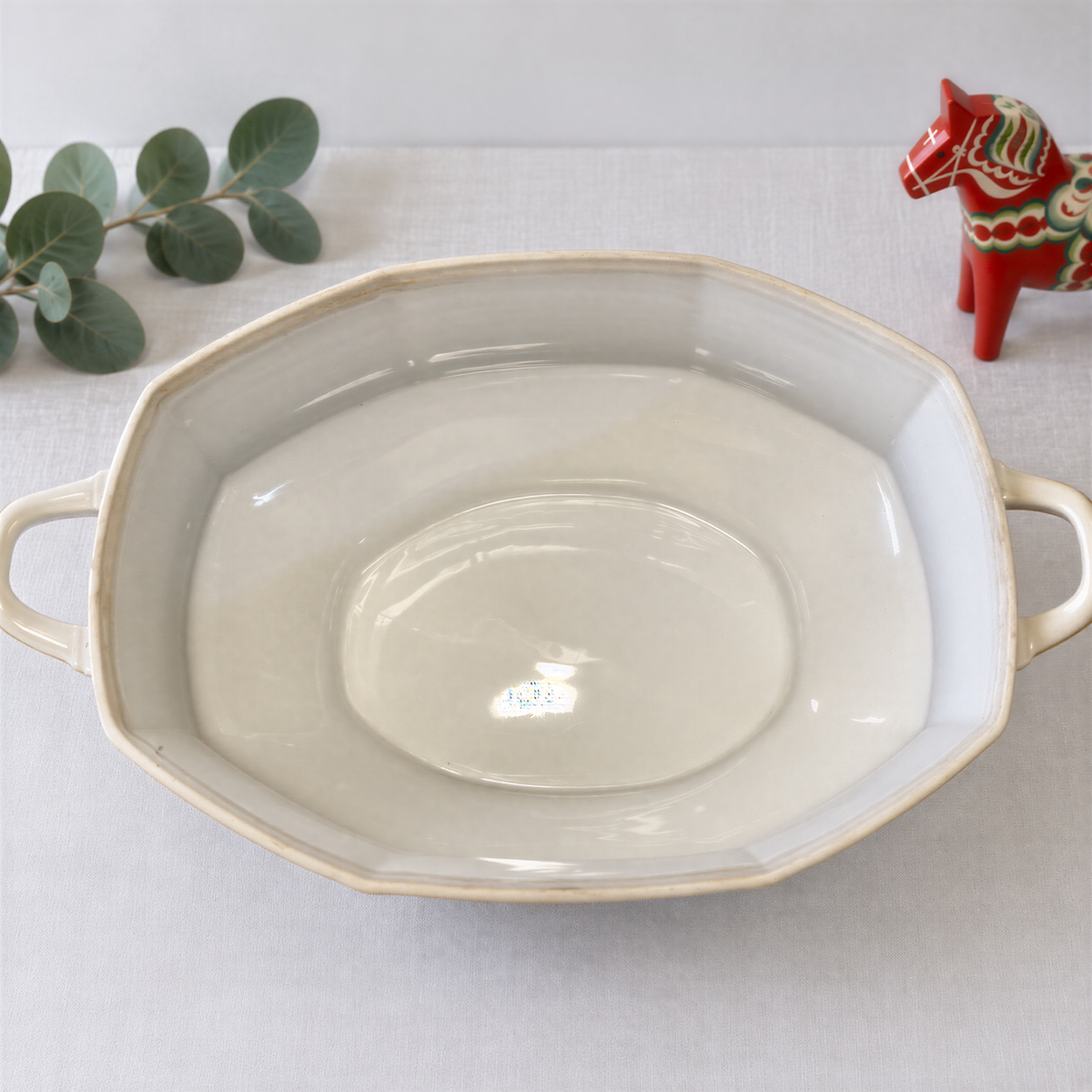 a white ceramic bowl with handles, placed on a white surface next to a small red and green ceramic figurine.