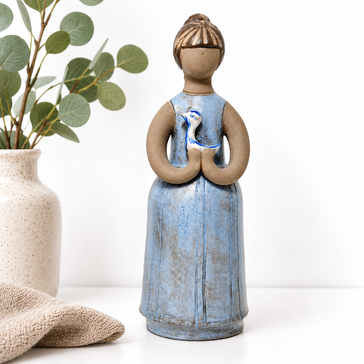 A ceramic figurine of a woman in a blue dress holding a bird is displayed on a white surface next to a white vase with green leaves.