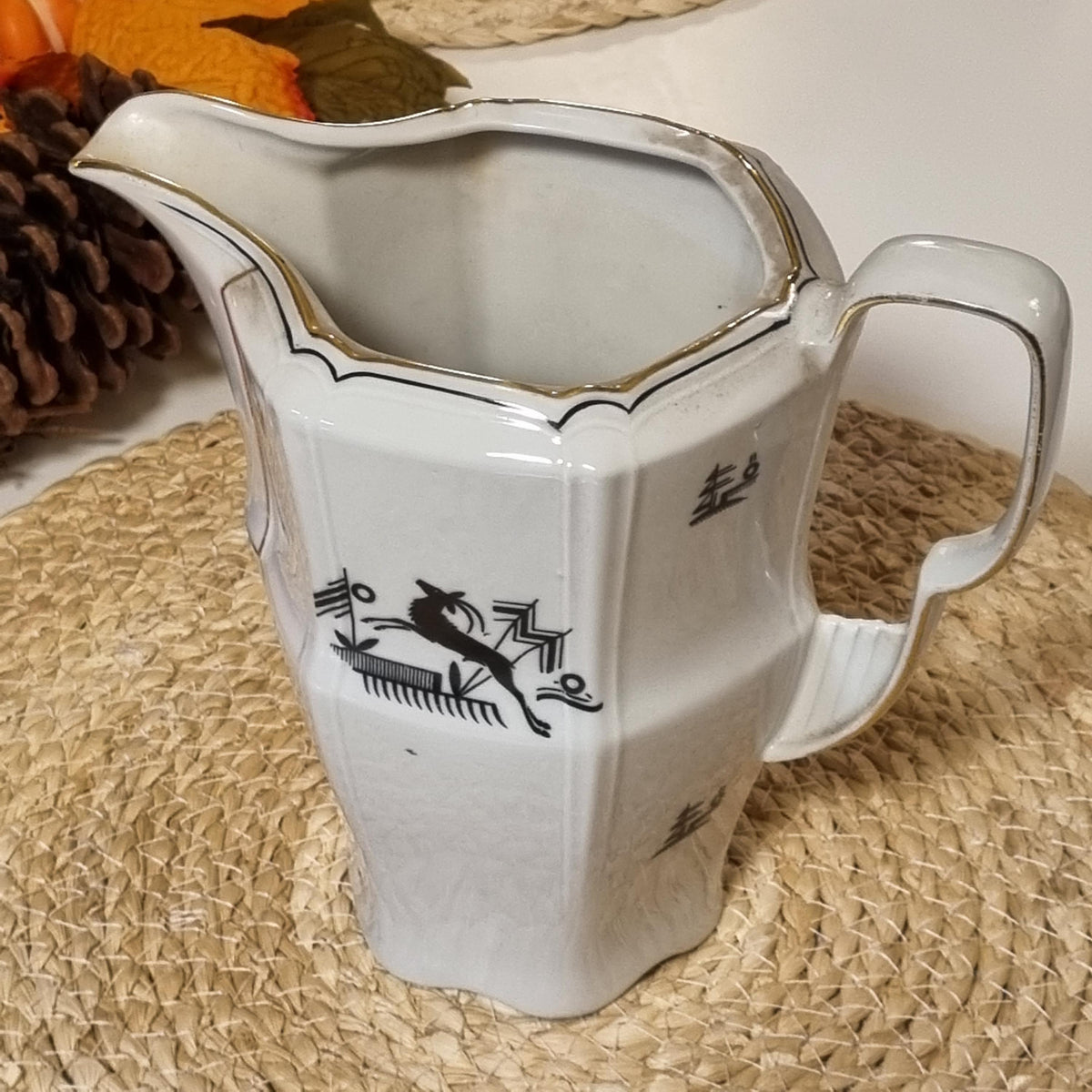 a white ceramic pitcher with a black design on it, placed on a woven surface.