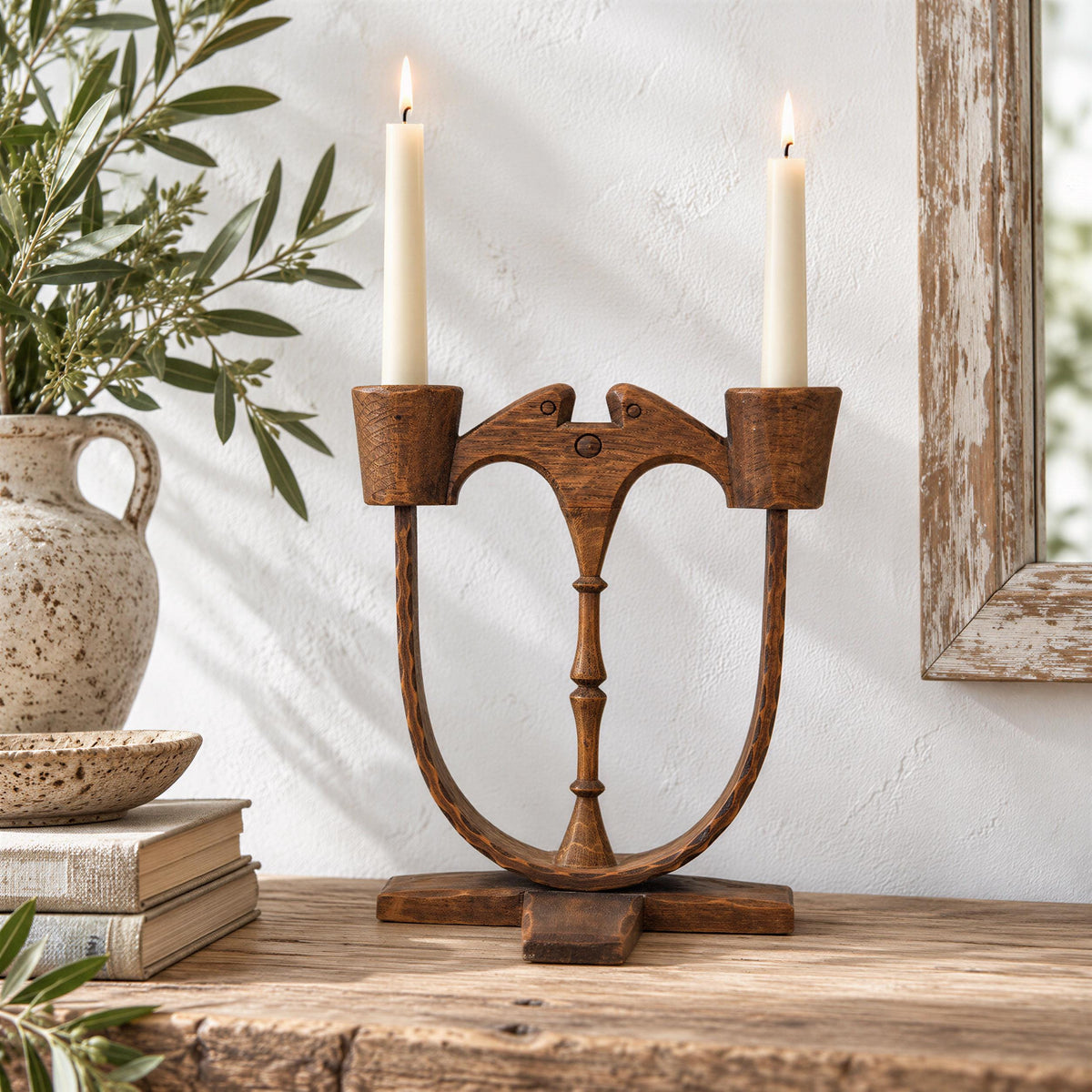 A rustic wooden candlestick holder with two white candles stands on a wooden surface, accompanied by a vase of greenery and a stack of books.