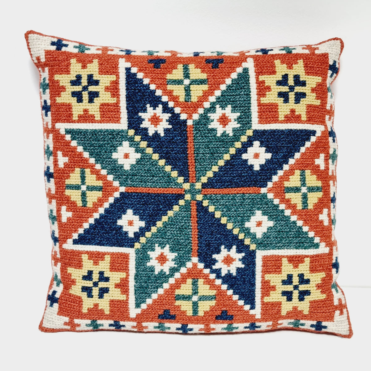 Square embroidered pillow with an eight-point star in navy and teal, framed by rust orange and cream motifs
