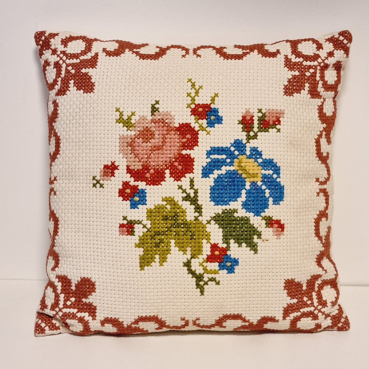 Vintage square cross-stitch pillow with a pink rose, blue flower and rust red ornamental border