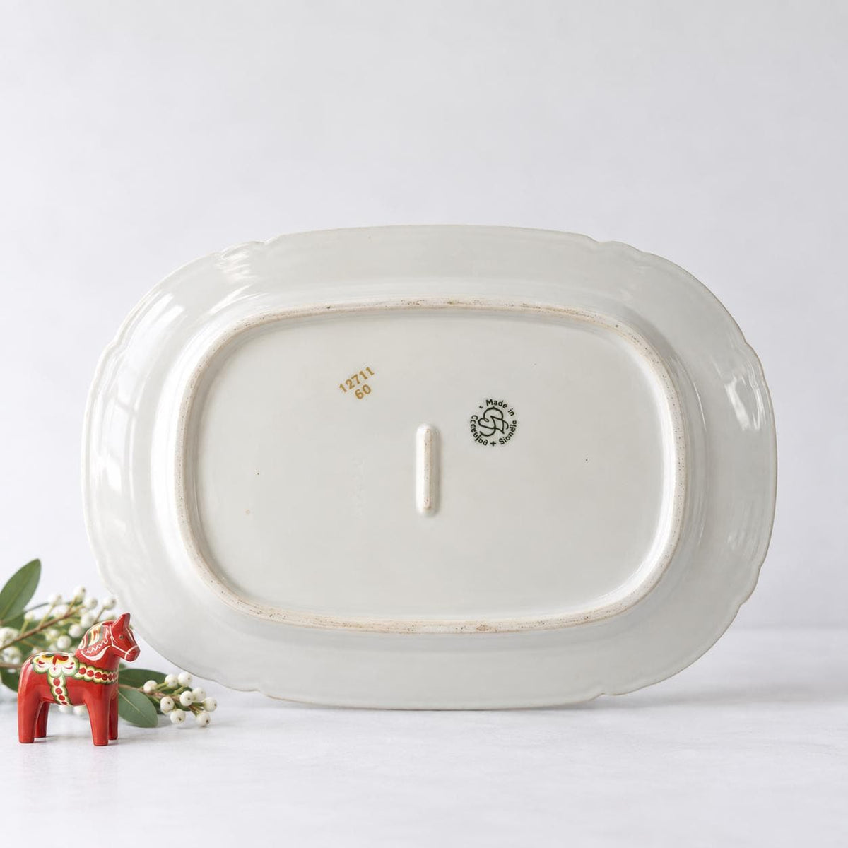 A white oval-shaped plate with a decorative border, placed on a white surface next to a small red figurine.