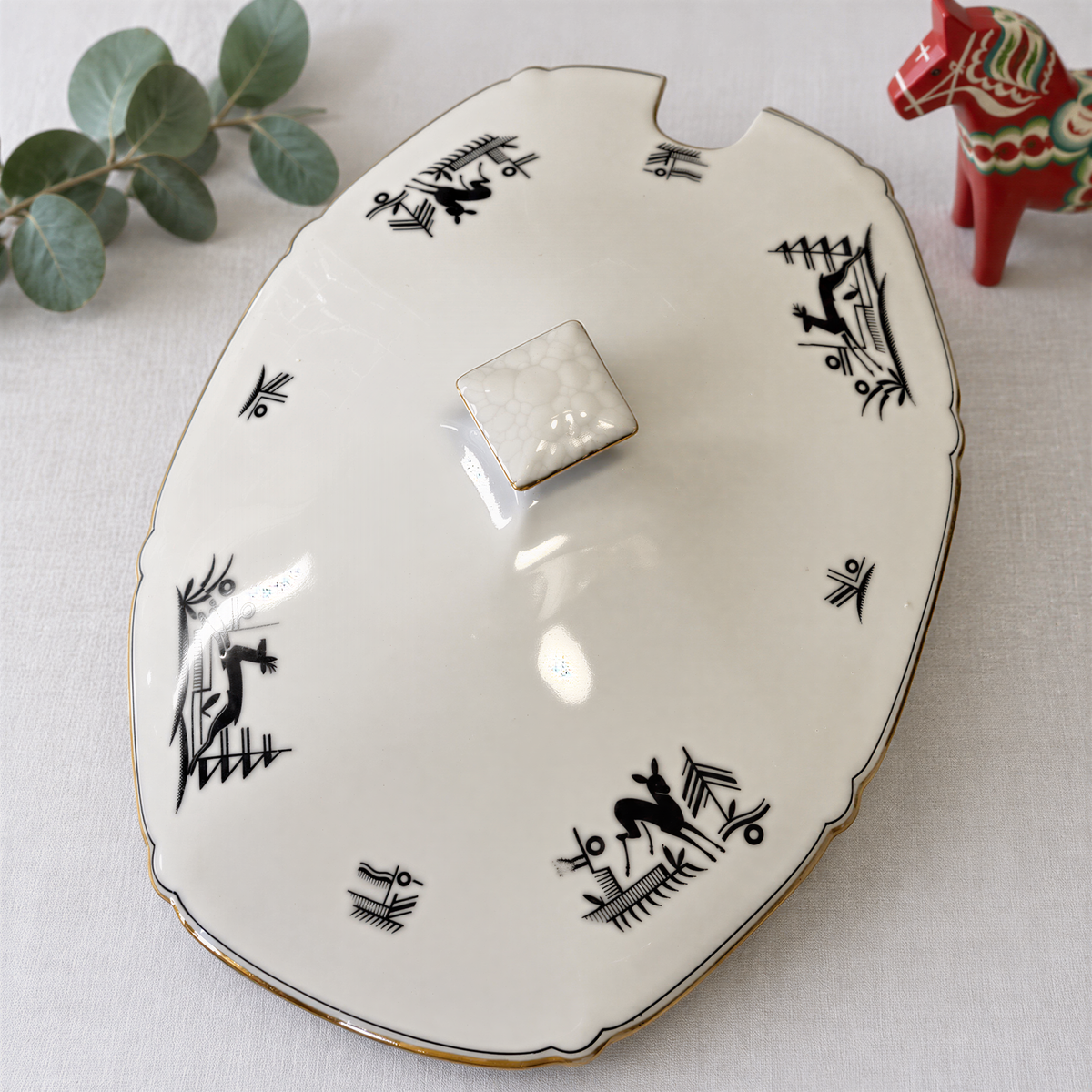 a white ceramic plate with black designs and a small white square piece on the top. There is also a small red figurine of a horse next to the plate.