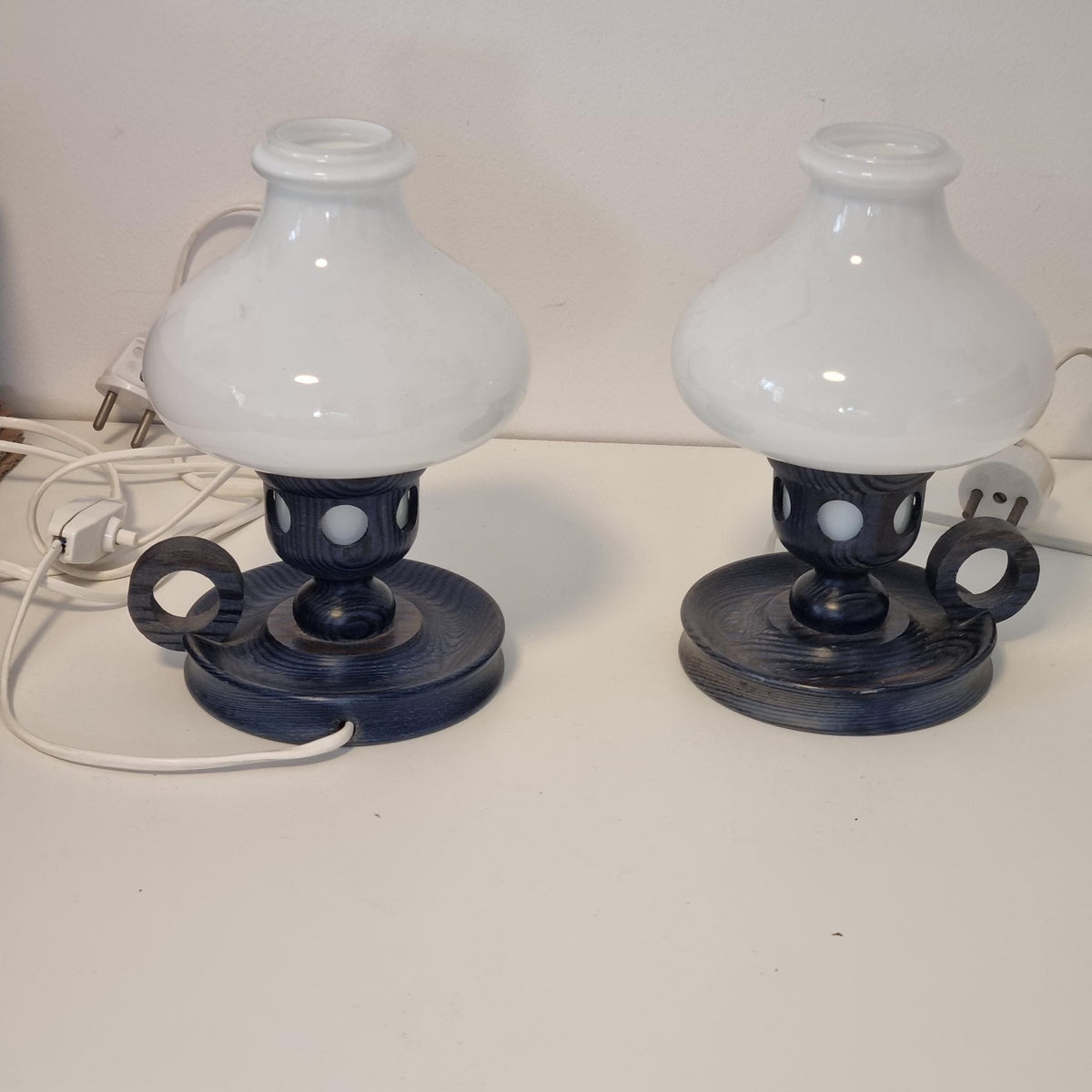 two wall-mounted lamps with white and black bases, each with a white shade. The lamps are connected to a white electrical outlet on the wall.