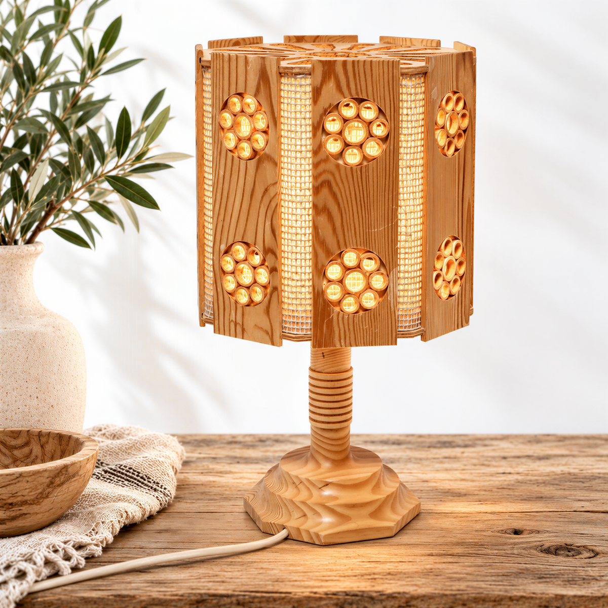 a wooden table lamp with a unique, circular design and a wooden base, placed on a wooden surface next to a white vase with green leaves.