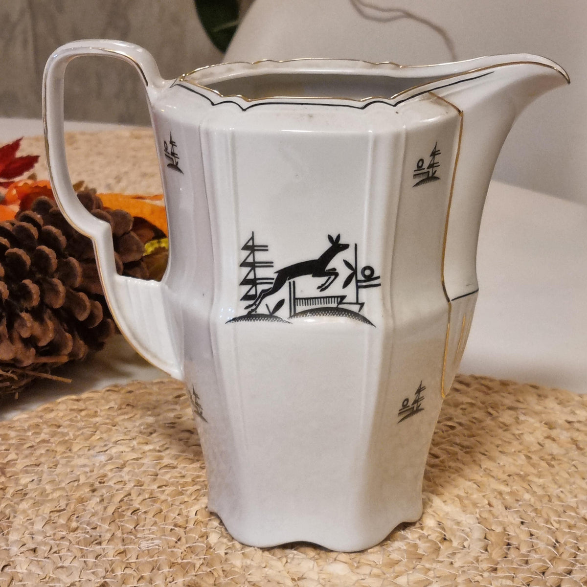 a white ceramic pitcher with a black and gold design on it, placed on a woven surface.