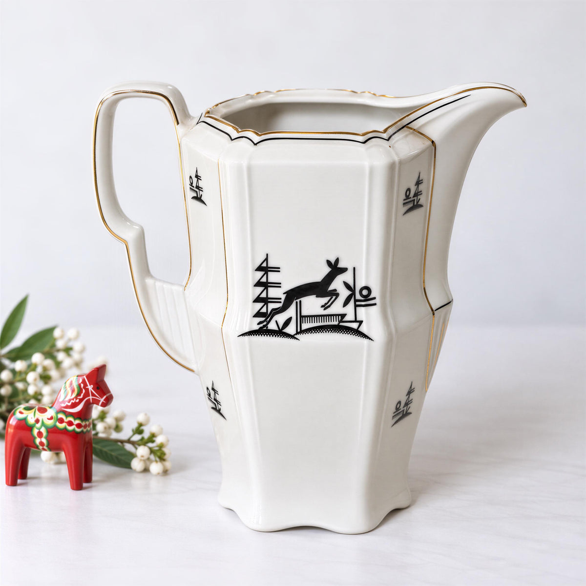 a white ceramic pitcher with a black and gold design, placed on a white surface next to a small red ceramic figurine.