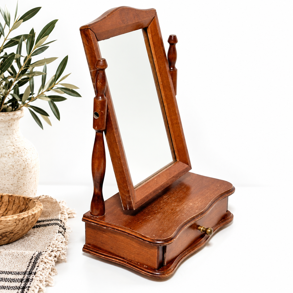 a wooden vanity with a mirror and a wooden drawer, placed on a white surface next to a vase with green leaves.
