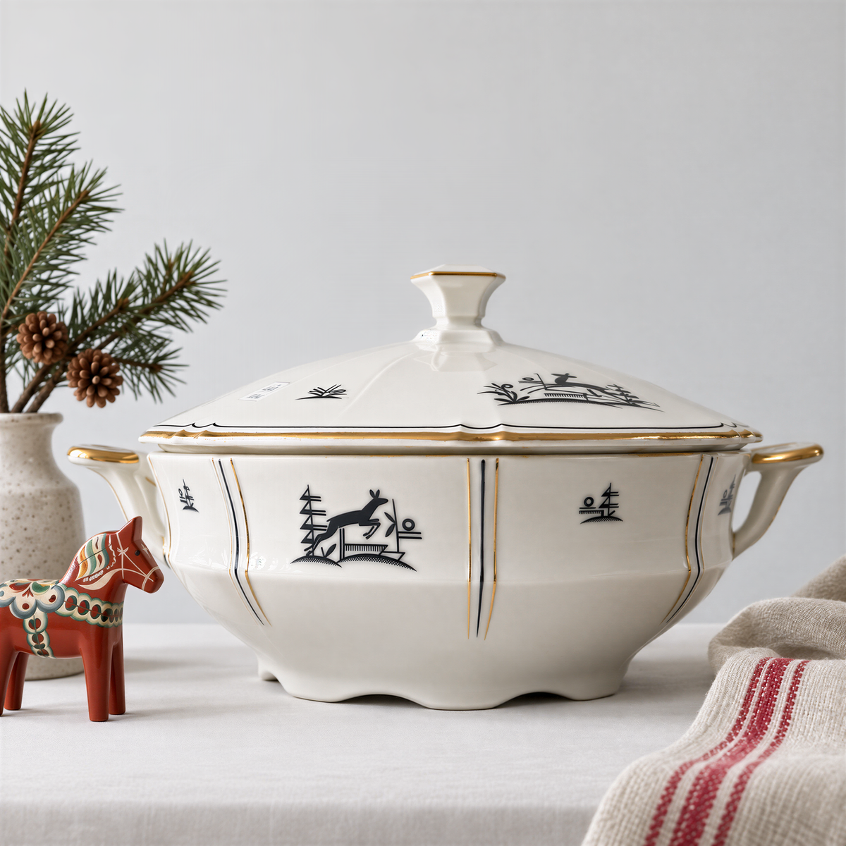 a white ceramic bowl with gold trim, decorated with black and gold designs, placed on a table next to a small ceramic figurine and a pine branch.