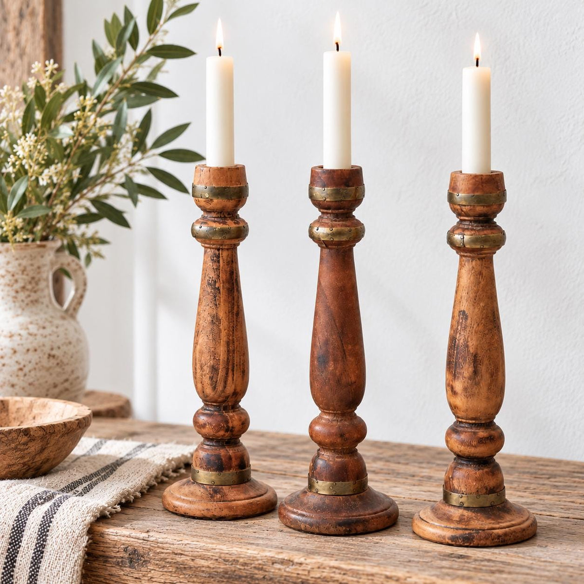 Three wooden candle holders with lit candles are placed on a wooden surface, accompanied by a vase of flowers and a striped cloth.