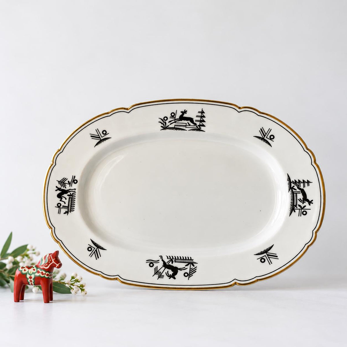 a white oval-shaped plate with black and gold designs, placed on a white surface next to a small red figurine.