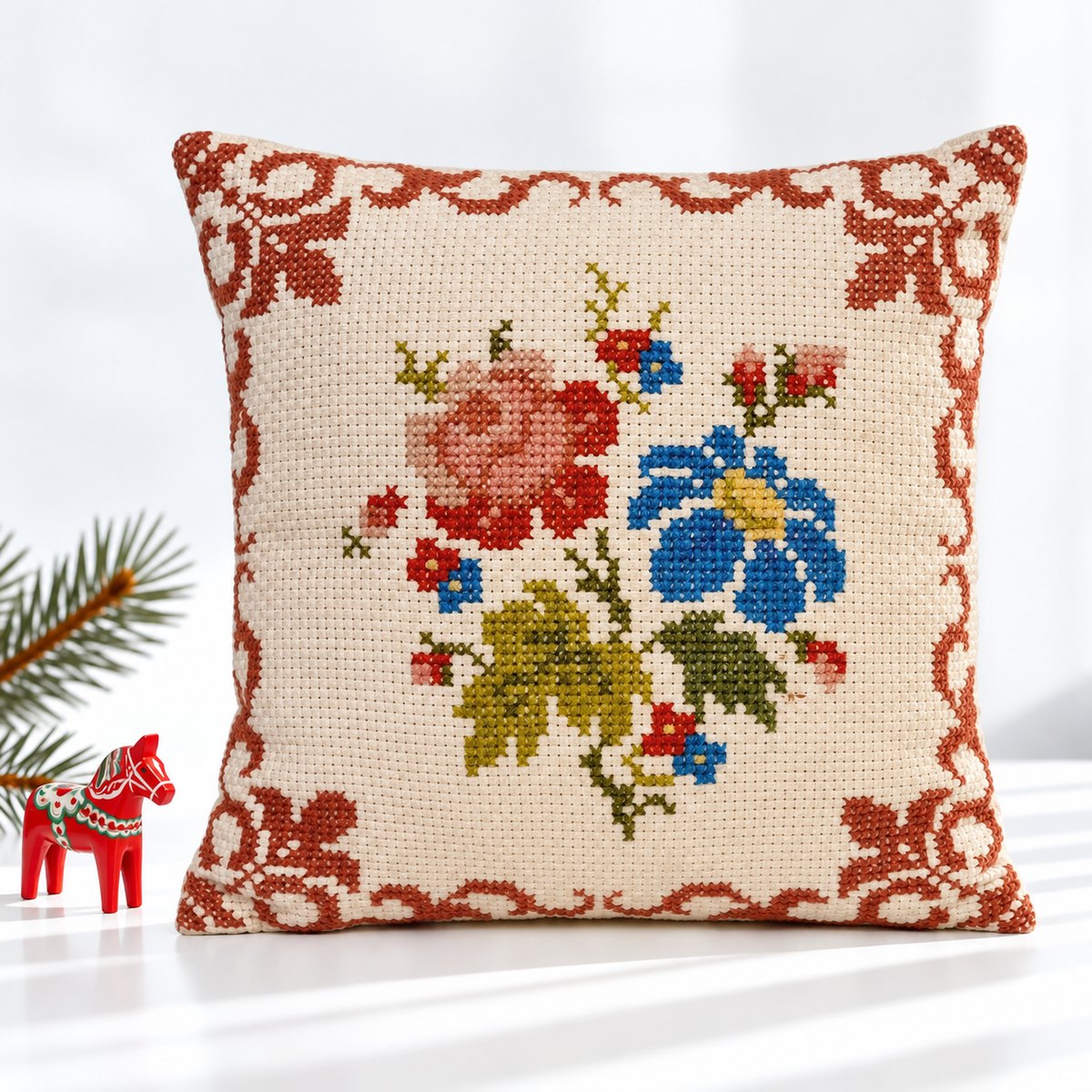 Floral cross-stitch pillow styled with a red Dala horse and evergreen branch
