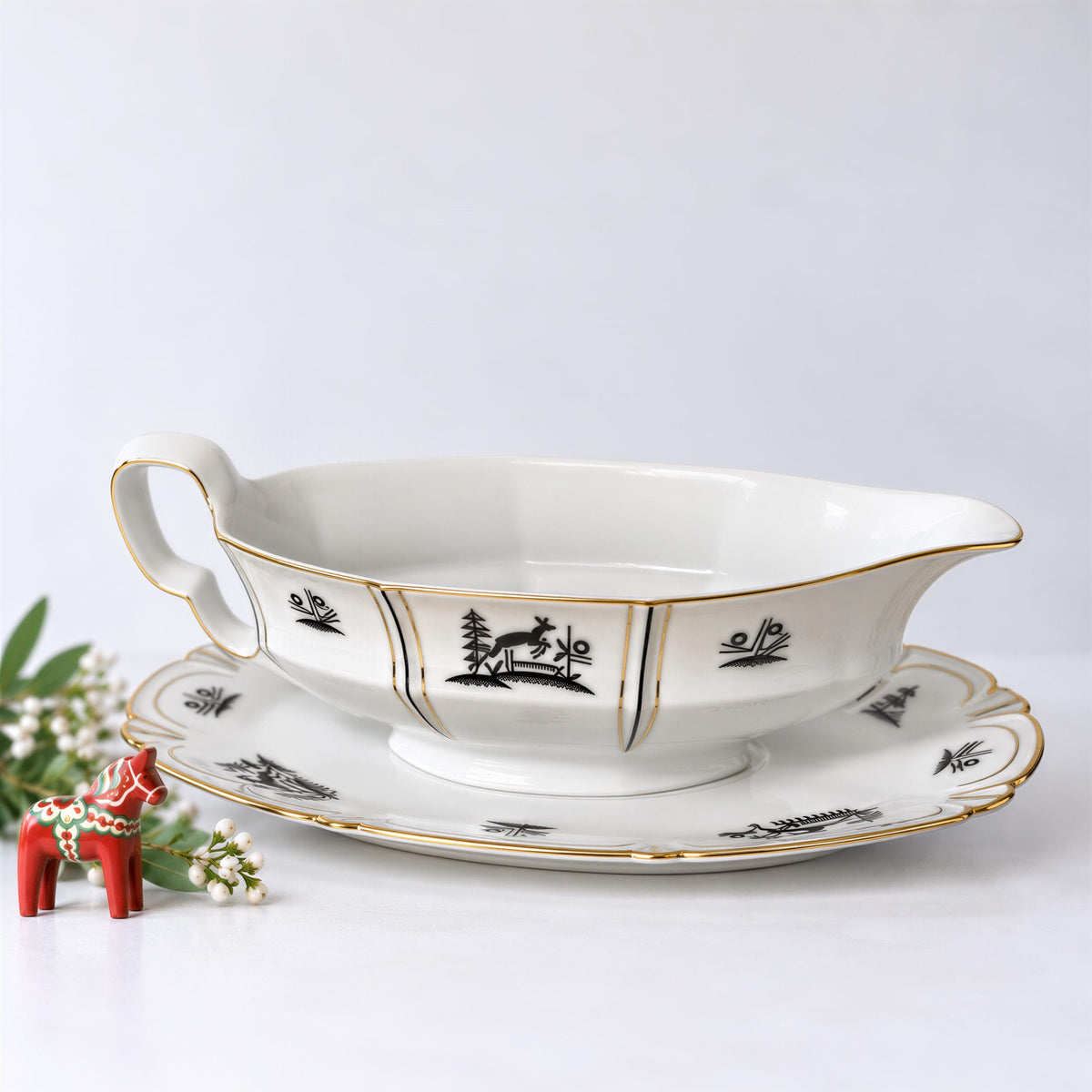 A white porcelain gravy boat with gold trim and black silhouettes of animals and trees sits on a white surface, accompanied by a small red figurine and some white flowers.