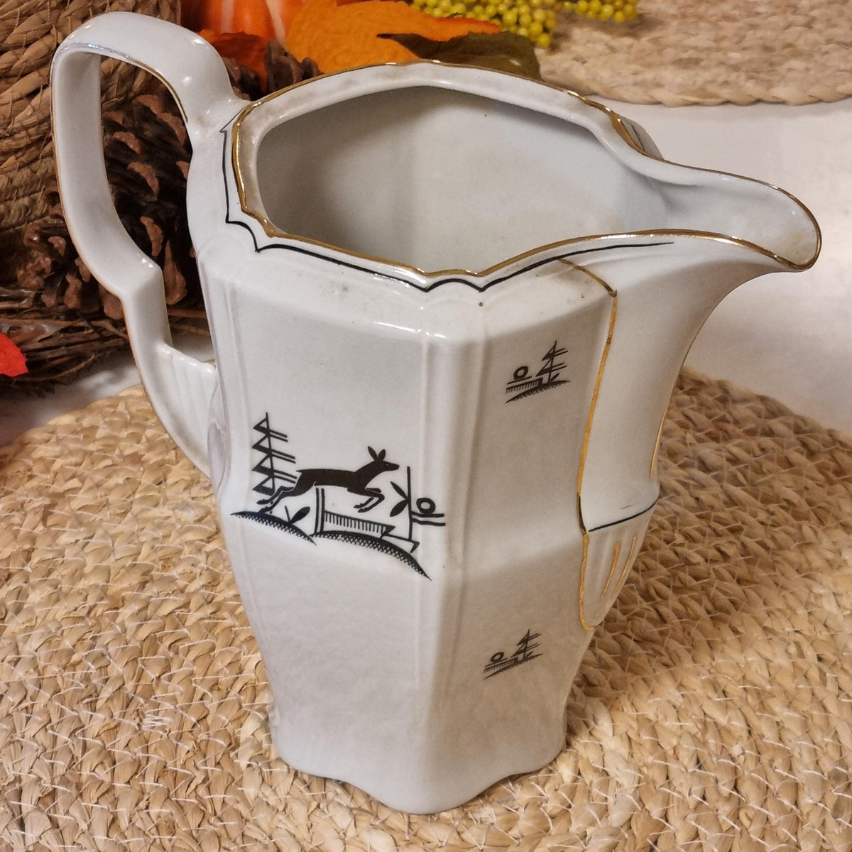 a white ceramic pitcher with a black and gold design on it, placed on a woven surface.