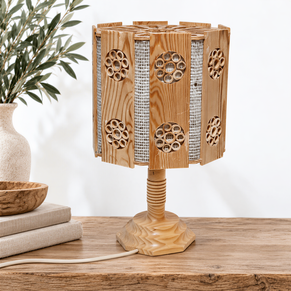 a wooden table lamp with a unique, circular design on its shade, placed on a wooden surface next to a stack of books and a vase with greenery.