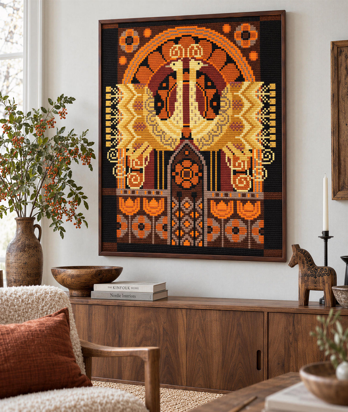 a large, colorful, and intricate tapestry hanging on a wall in a living room. The tapestry is composed of various geometric shapes and patterns in shades of orange, yellow, and black. The room is decorated with a wooden cabinet, a vase, a horse figurine, and several books, creating a warm and inviting atmosphere.