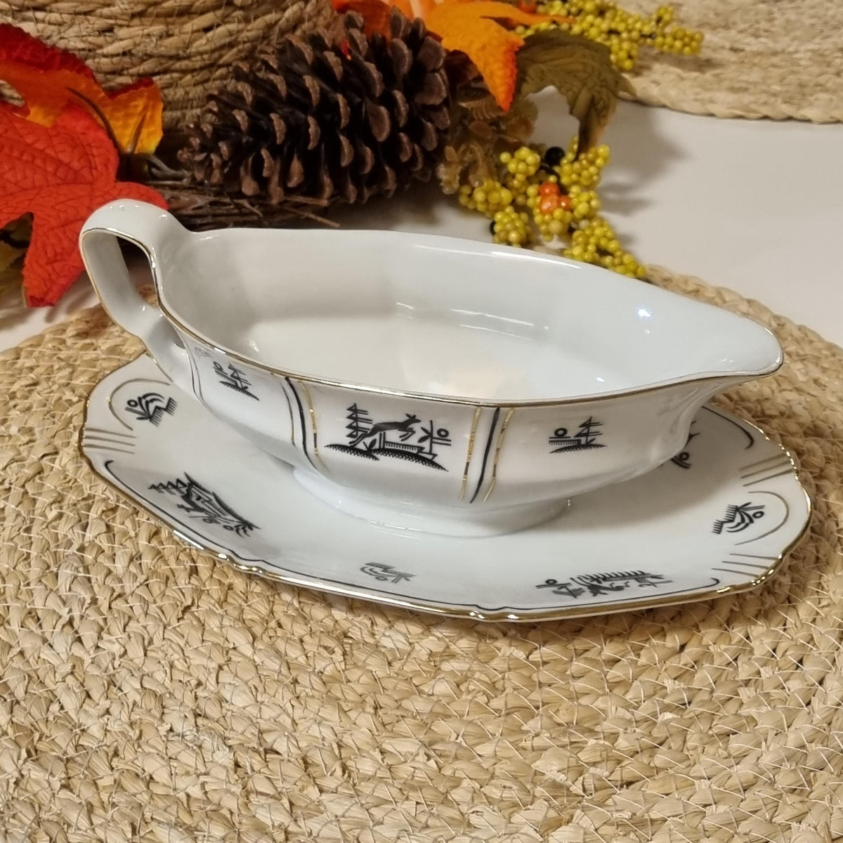 A white porcelain gravy boat and saucer with black decorative patterns are placed on a woven placemat, surrounded by autumn leaves and pine cones.