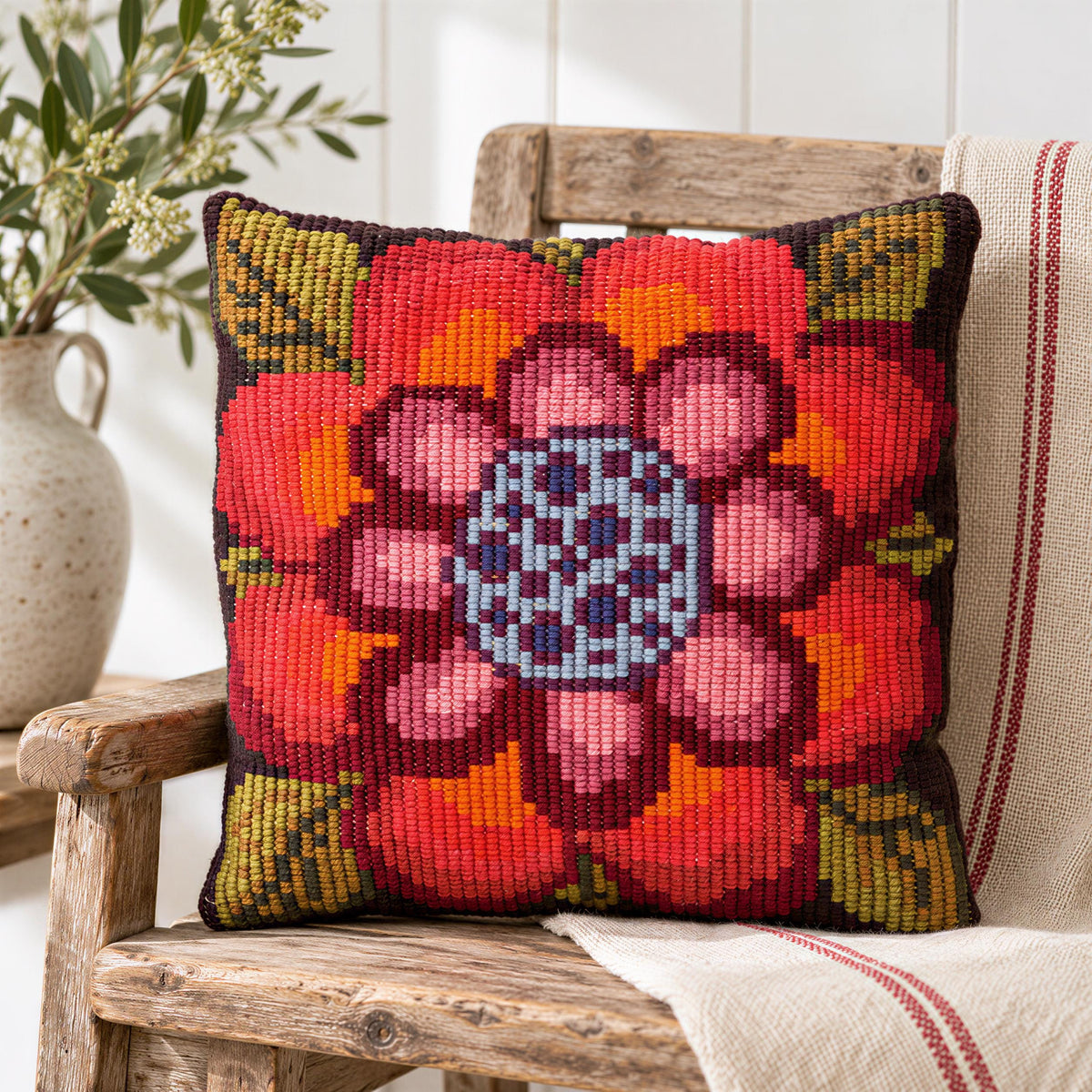 A vibrant, hand-knitted pillow with a large, colorful flower design sits on a wooden chair, with a vase of flowers and a striped towel in the background.