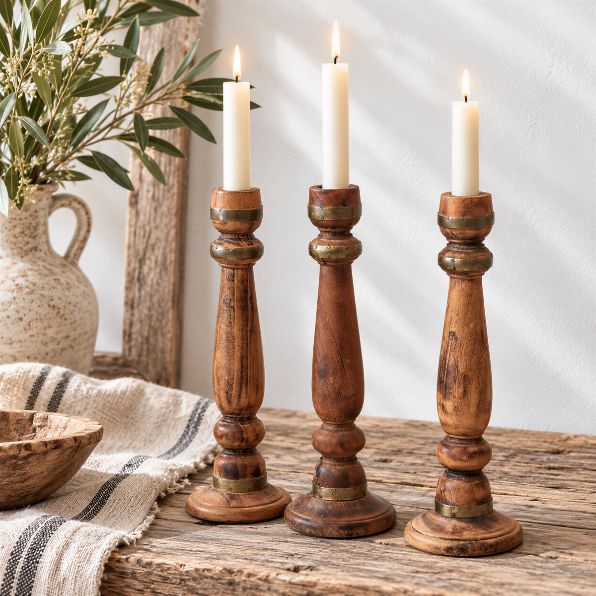three wooden candle holders with white candles, placed on a wooden surface. There is a vase with greenery and a striped cloth in the background.