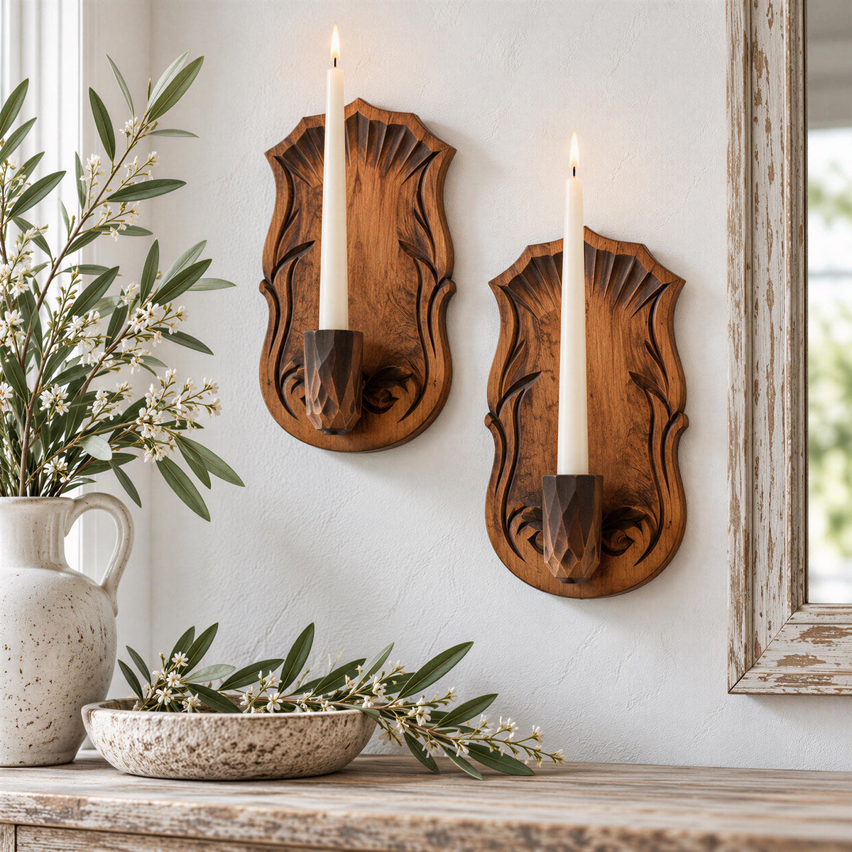 a rustic wooden candle holder mounted on a white wall, with two white candles burning inside. The candle holder has an ornate design, adding a touch of elegance to the decor. In the foreground, there is a white vase filled with greenery, and a bowl containing white flowers. The arrangement creates a warm and inviting atmosphere in the room.